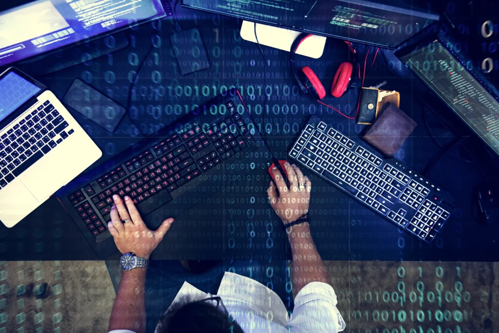 Mão branca masculina digitando no teclado enquanto trabalha com programação. Está sentado em uma mesa preta de frente para três monitores e um macbook.